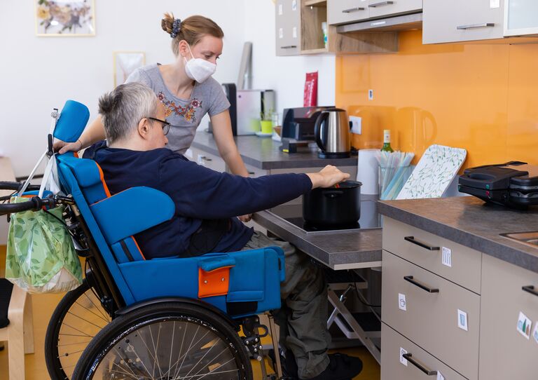 Ein Kunde im Rollstuhl wird von einer Mitarbeiterin zum Schreibtisch geschoben.