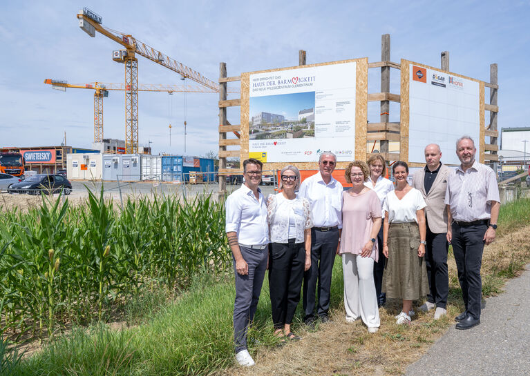 Sozial-Landesrätin Christiane Teschl-Hofmeister, Bürgermeister Johannes Hölzl sowie Vertreter:innen des Haus der Barmherzigkeit vor einer Bautafel und Abschnitt der Baustelle.