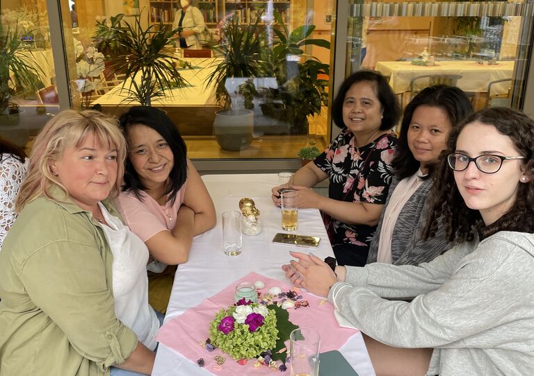 Kolleg*innen sitzen am Esstisch outdoor zusammen und lachen in die Kamera.