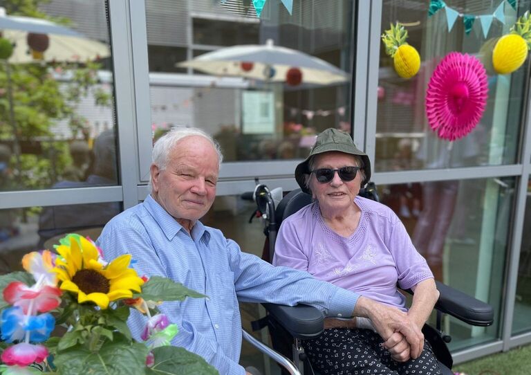 Eine Bewohnerin im Rollstuhl und neben ihr sitzt ihr Ehemann, der seine Hand auf ihren Schoß legt.