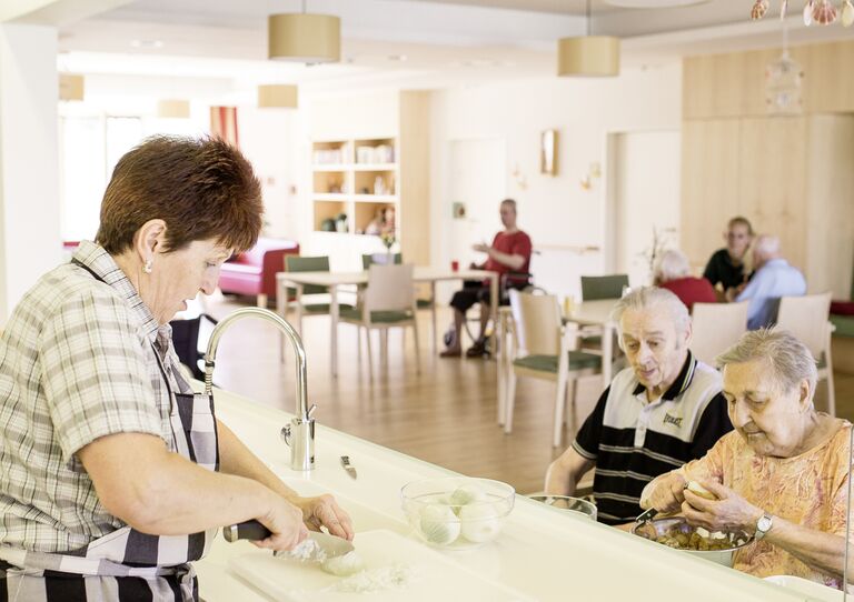 Eine Betreuerin bereitet in der gemeinsamen Küche einer Hausgemeinschaft das Essen zu. Zwei Bewohnerinnen helfen dabei.