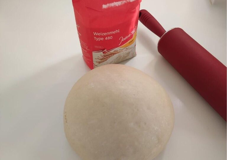 Germteig im rohen Zustand zusammengerollt zu einem Ball, dahinter steht ein Nudelwalger und ein Sack Mehl.