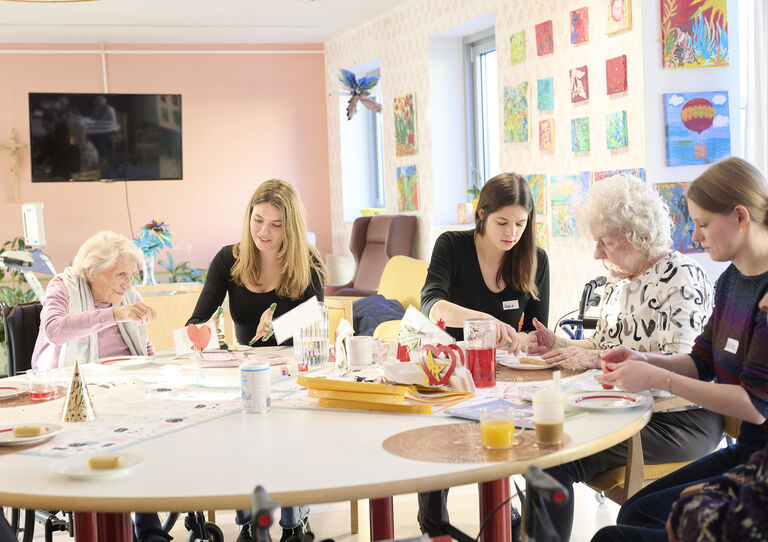 Studierende absolvieren das Praktikums. Sie sitzen gemeinsam mit Bewohner*innen um einen Tisch und führen Gespräche beim Basteln.  Studierende absolvieren das Praktikums. Sie sitzen gemeinsam mit Bewohner*innen um einen Tisch und führen Gespräche beim Basteln.