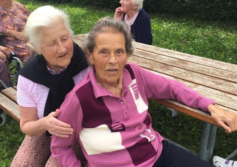 Zwei Bewohnerinnen sitzen auf Bank und halten einander ein.