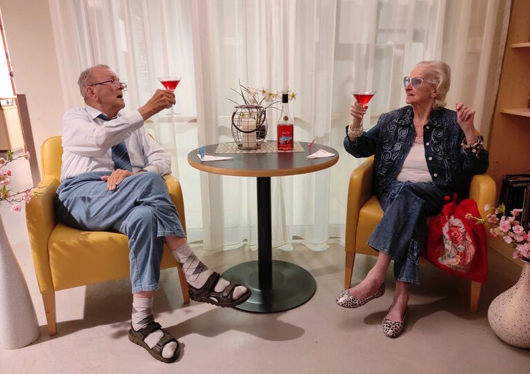 Eine Bewohnerin und ein Bewohner sitzen sich an einem Kaffeehaustisch gegenüber und prosten sich mit einem pinken Prosecco zu.