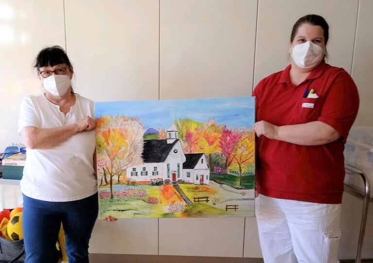 Gabi Posch und eine Mitarbeiterin halten ein Kunstwerk, das ein Haus im Grünen zeigt.