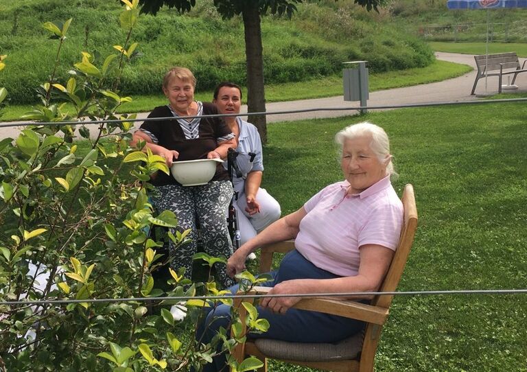Zwei Bewohnerinnen sitzen neben dem Strauch und lächeln in die Kamera und hinter ihnen sitzt Alltagsbetreuerin Marija.