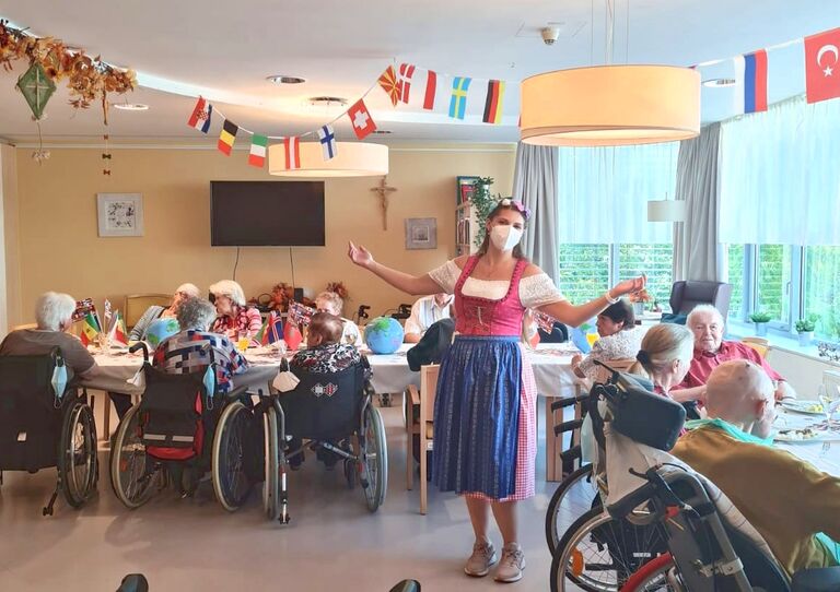 Stationsleiterin Carina Thallinger steht im Aufenthaltsraum der St. Christoph und präsentiert die schöne Dekoration. Hinter ihr sitzen die Bewohner*innen an den Tischen und essen vom köstlichen Buffet.