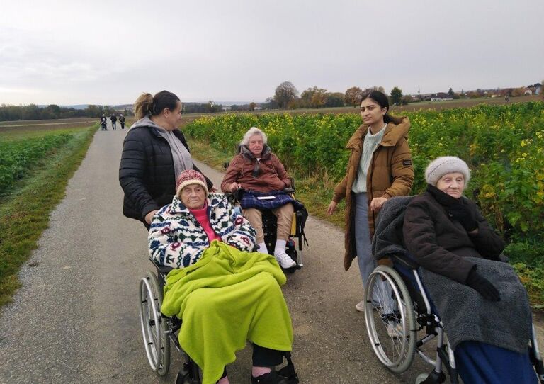Bewohner*innen im Rollstuhl mit Decke. Hinter dem Rollstuhl stehen die Mitarbeiter*innen.