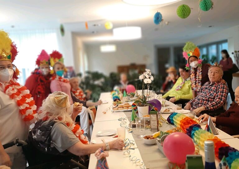 Bewohnerinnen sitzen an einem bunt dekorierten Tisch. Mitarbeiter*innen stehen dahinter.